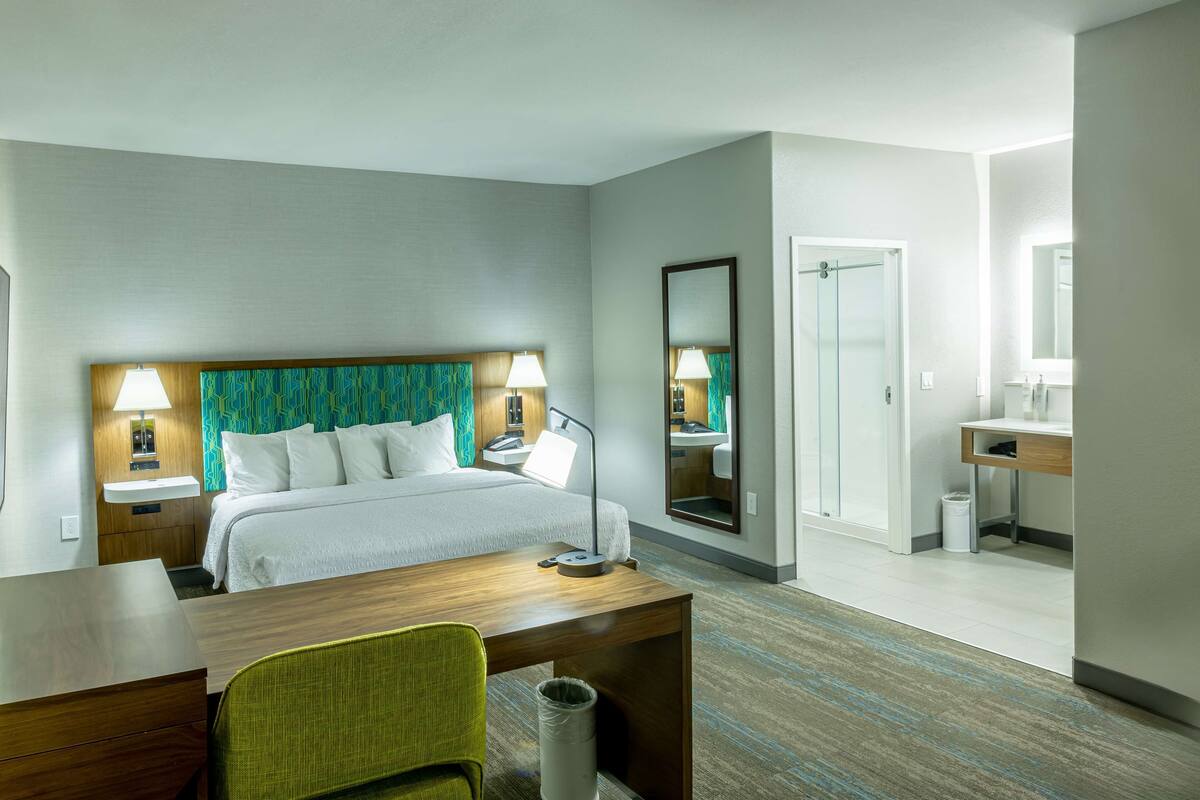 A hotel room with a bed, bedside tables, a desk, a chair, a mirror, a shower, and a sink.