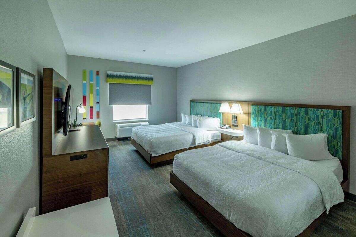 A hotel room with two beds, a desk, a TV, and a window.