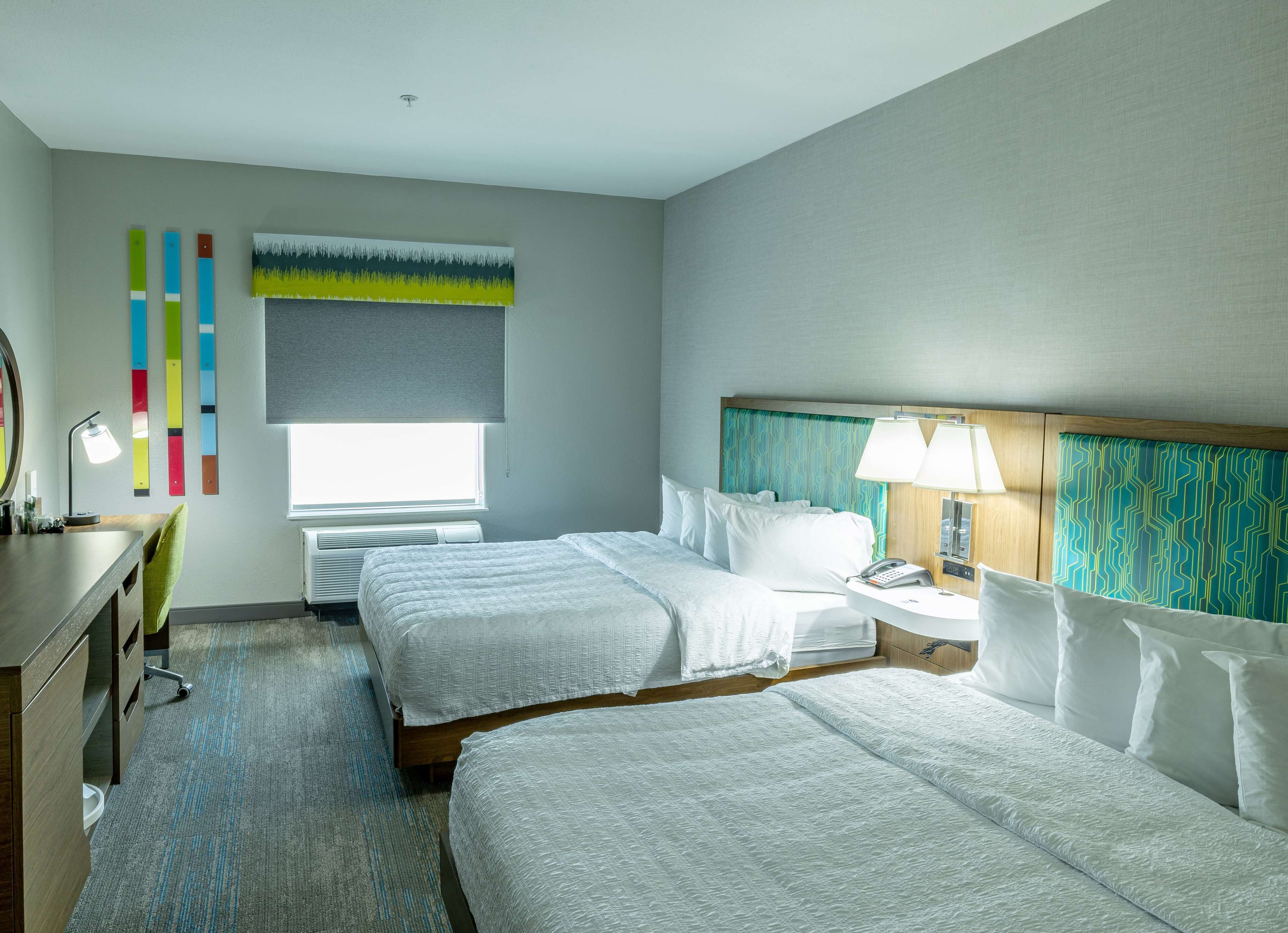 Habitación de hotel con dos camas, un escritorio, una silla, un ventanal con vistas y una pared con decoración colorida.