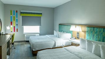 Habitación de hotel con dos camas, un escritorio, una silla, un ventanal con vistas y una pared con decoración colorida.