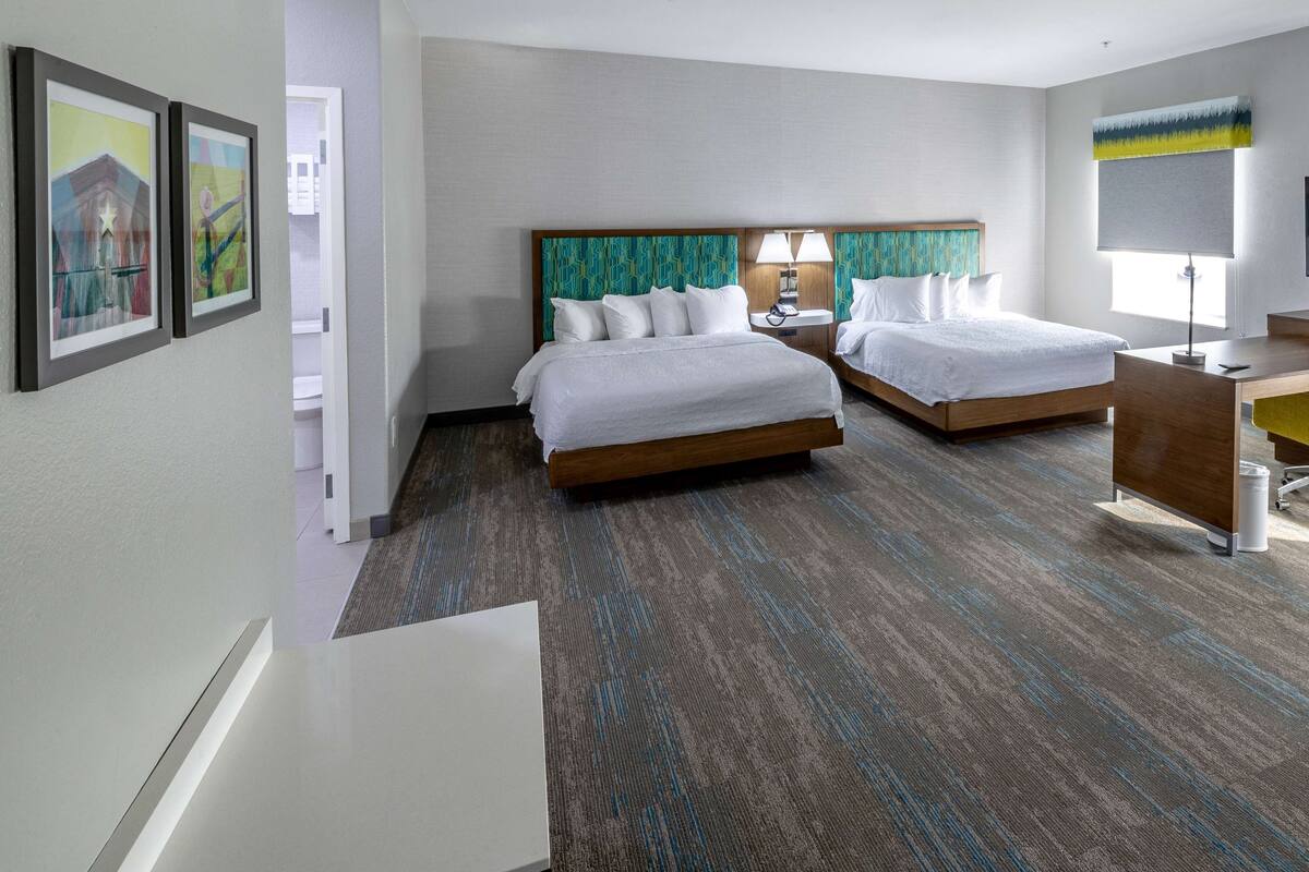 A hotel room with two beds, a desk, a chair, a lamp, and framed artwork on the wall.