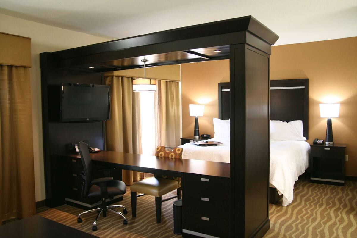 A hotel room with a desk, chair, bed, bedside tables, lamps, and a TV.
