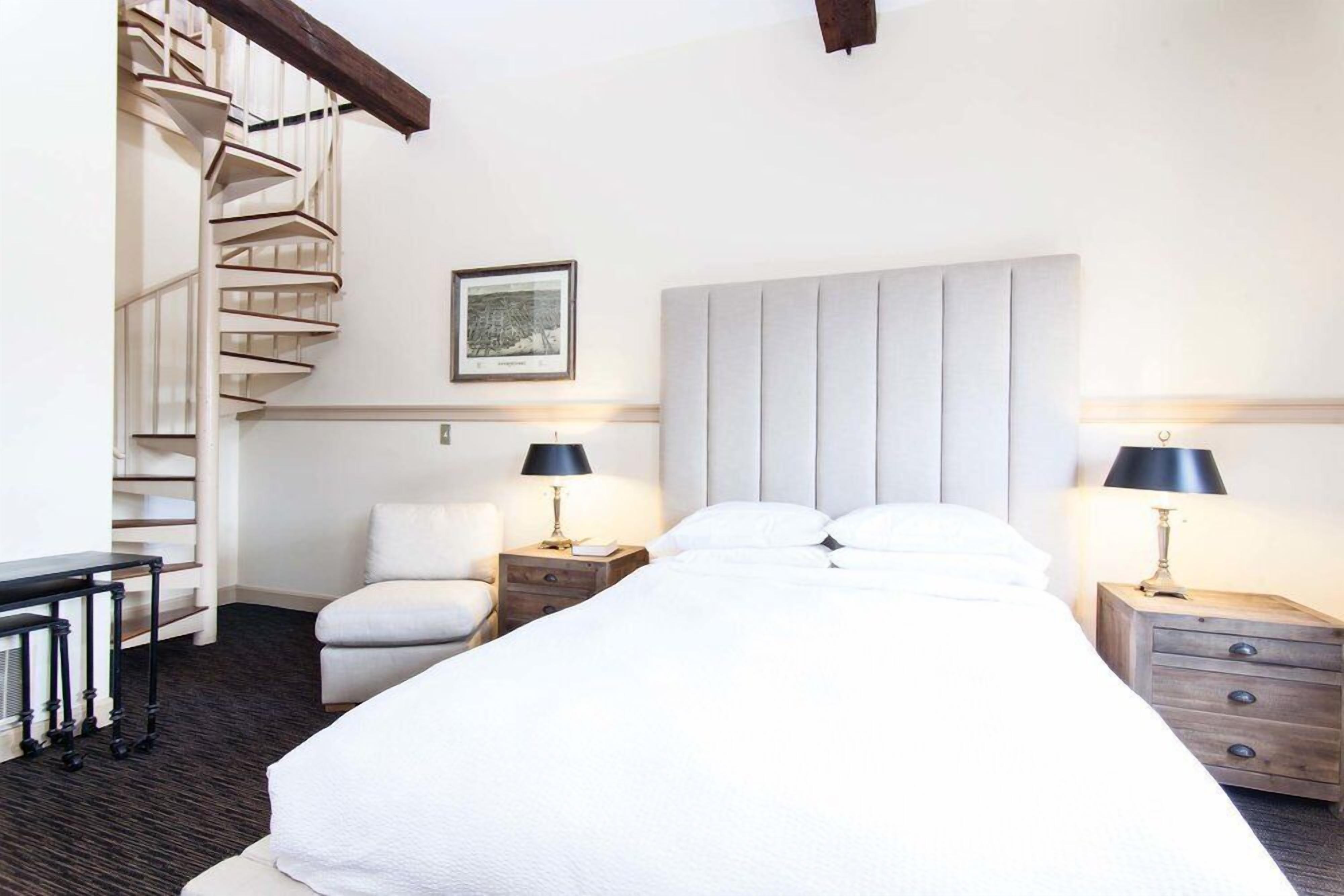 A hotel room with a bed, two bedside tables, a chair, a desk, and a spiral staircase.