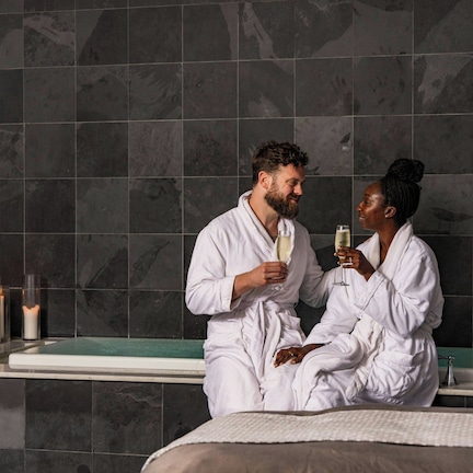 A couple in white robes sitting in a bathtub with candles and a bottle of champagne.