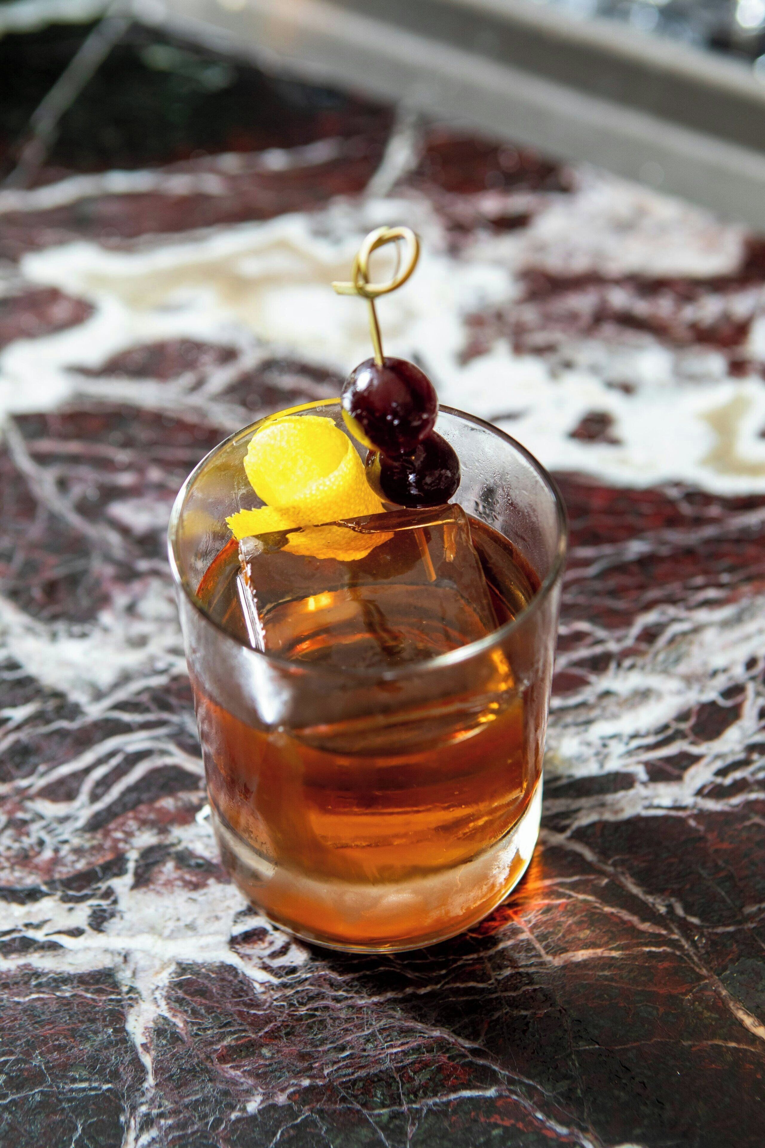 A glass of amber-colored beverage with a cherry and orange slice garnish on a textured surface.