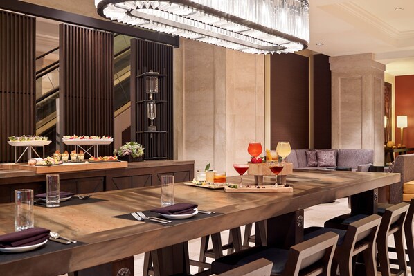 A modern dining area with a long wooden table, bar stools, and a bar counter with a wine cooler. There is a seating area with a sofa and a lamp.