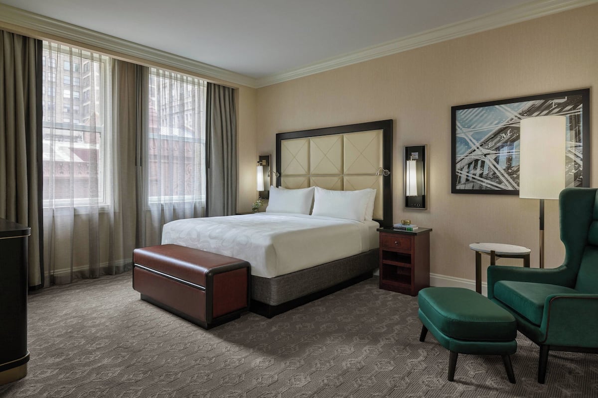 Une chambre d’hôtel avec un grand lit, un fauteuil vert, une petite table et une grande fenêtre avec des rideaux.
