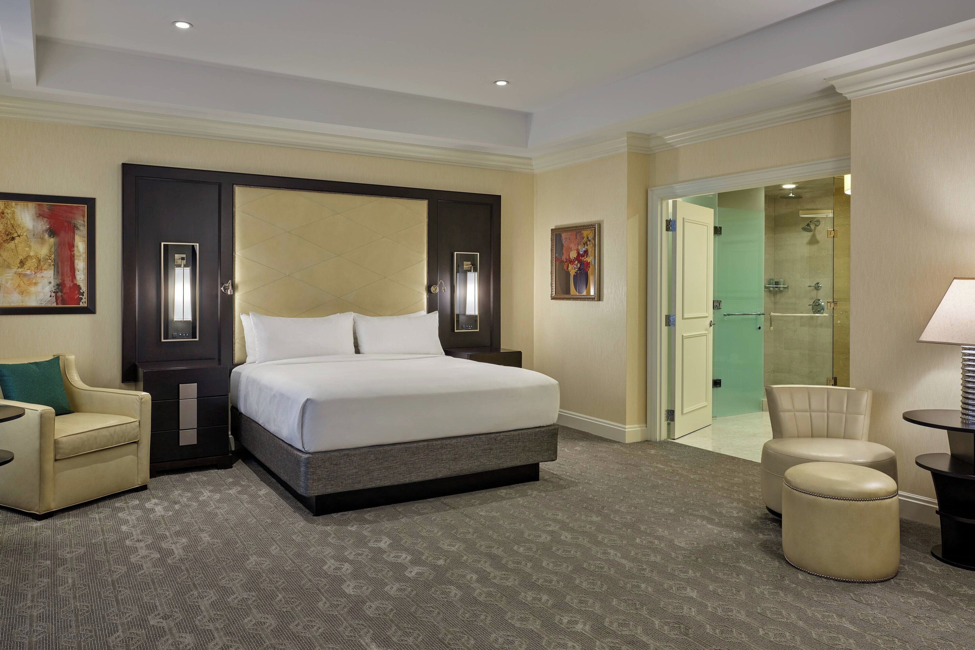 Une chambre d’hôtel avec un grand lit, un coin salon comprenant un fauteuil, une petite table, un miroir et une porte donnant sur une salle de bain.