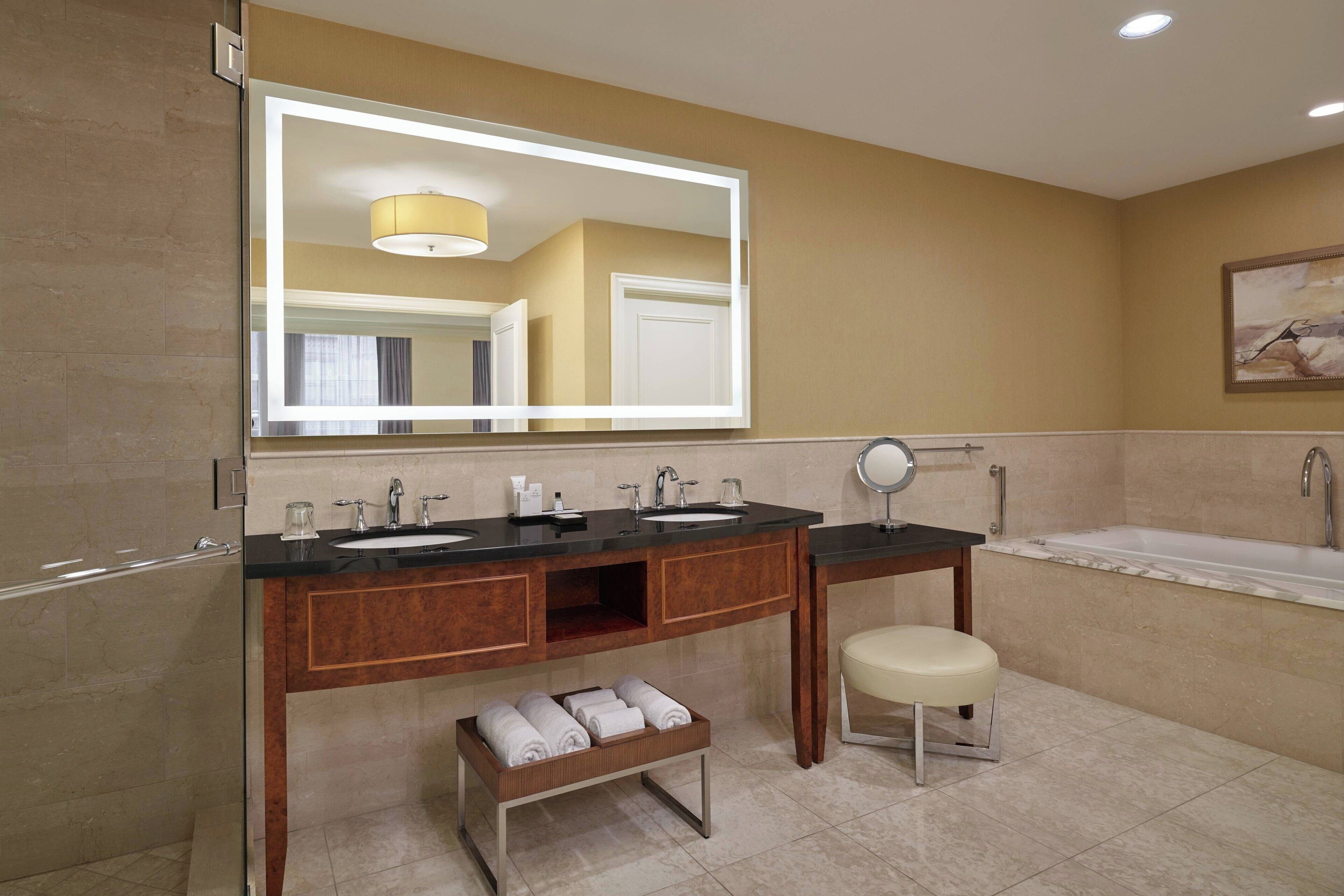 A modern bathroom with two sinks, a vanity, a bench with towels, a large mirror, and a bathtub.