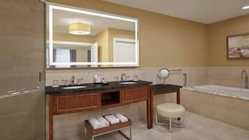 A modern bathroom with two sinks, a vanity, a bench with towels, a large mirror, and a bathtub.
