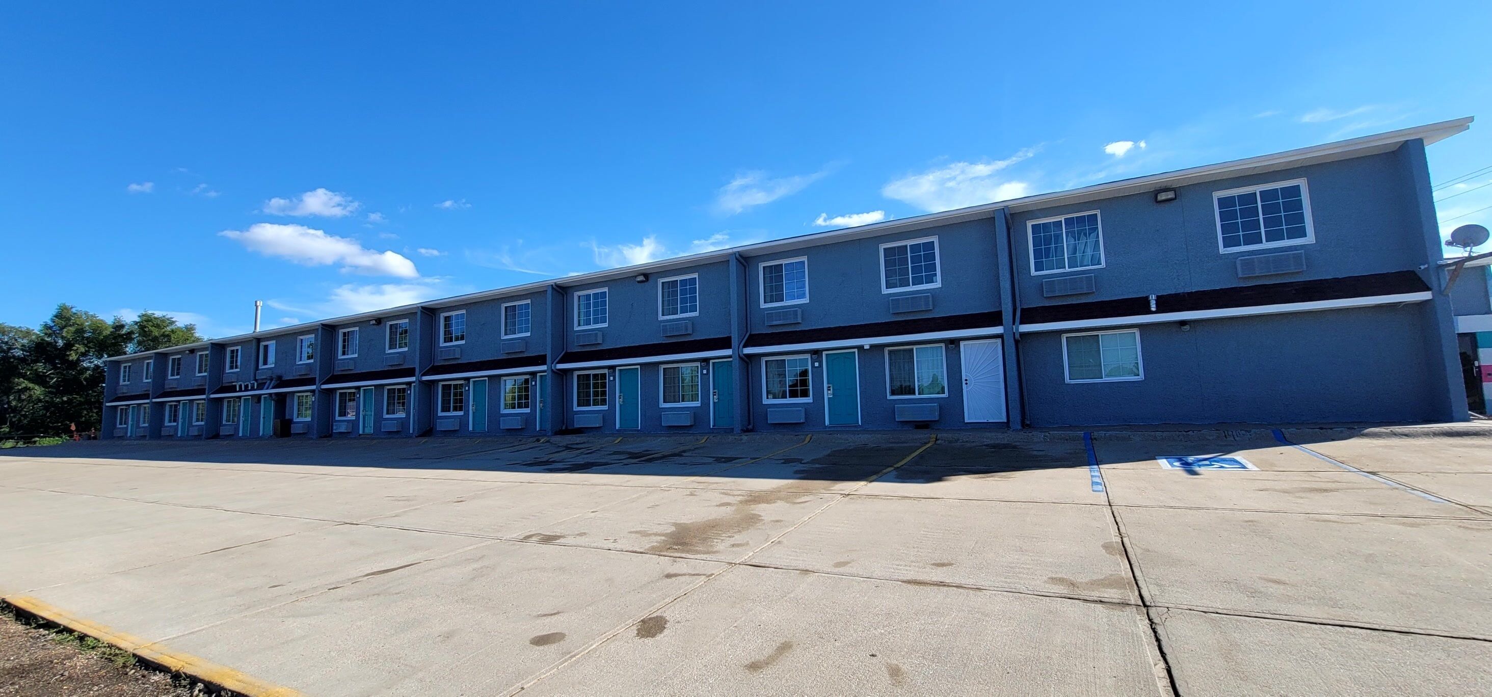 Um motel com vários andares, uma fileira de quartos, um estacionamento e um céu azul claro.