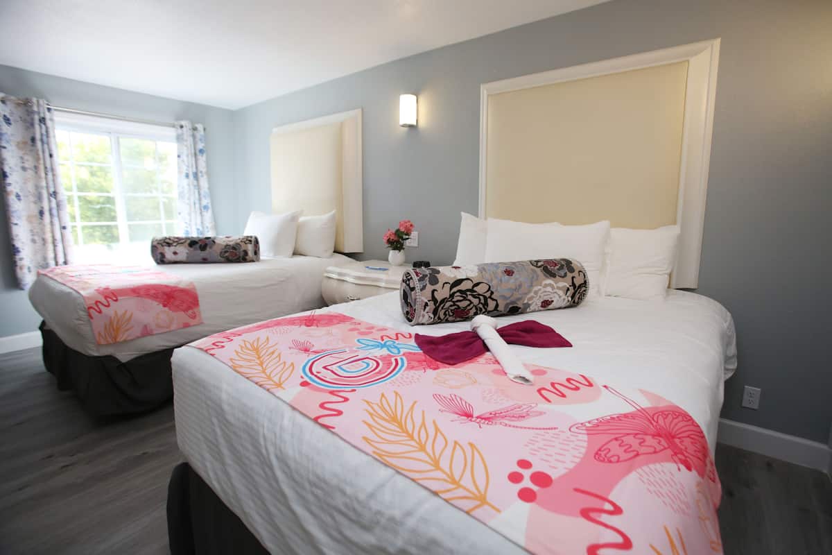 A hotel room with two beds, each with a distinct floral bedspread. There is a window with curtains, a bedside table with a vase of flowers, and a wall-mounted light.