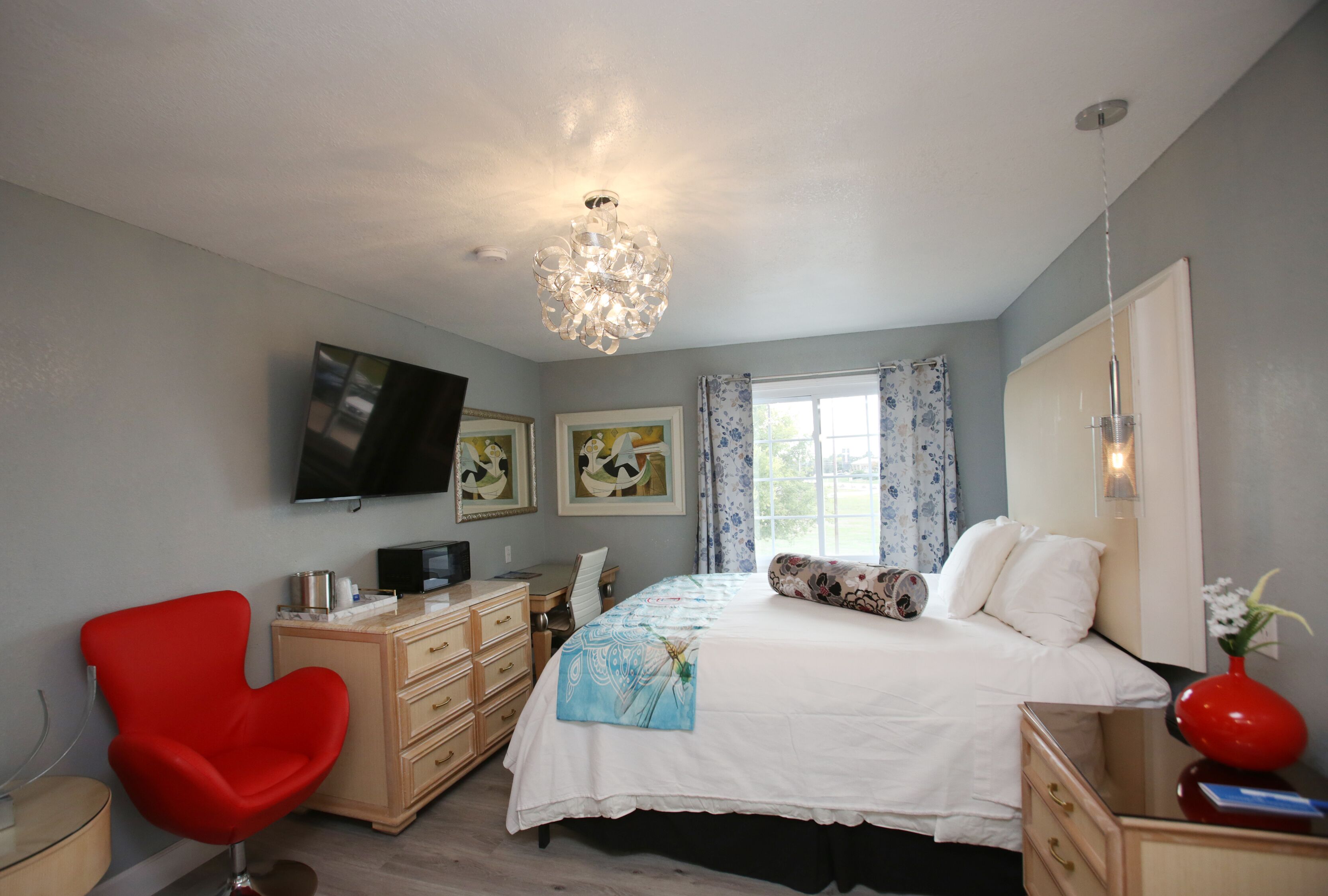 A bedroom with a bed, a red chair, a dresser, a television, and artwork on the wall.