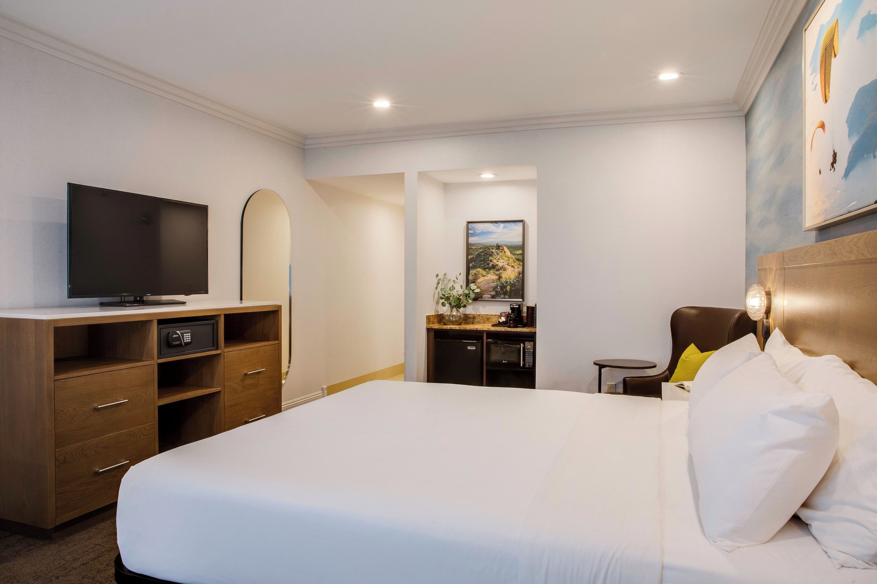 A hotel room with a large bed, a TV, a desk with a computer, a small kitchen area, and a painting on the wall.
