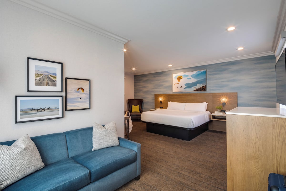 Ein modernes Hotelzimmer mit einem blauen Sofa, einem Bett mit weißer Bettwäsche, einem Holztisch und einem an der Wand befestigten Fernseher.