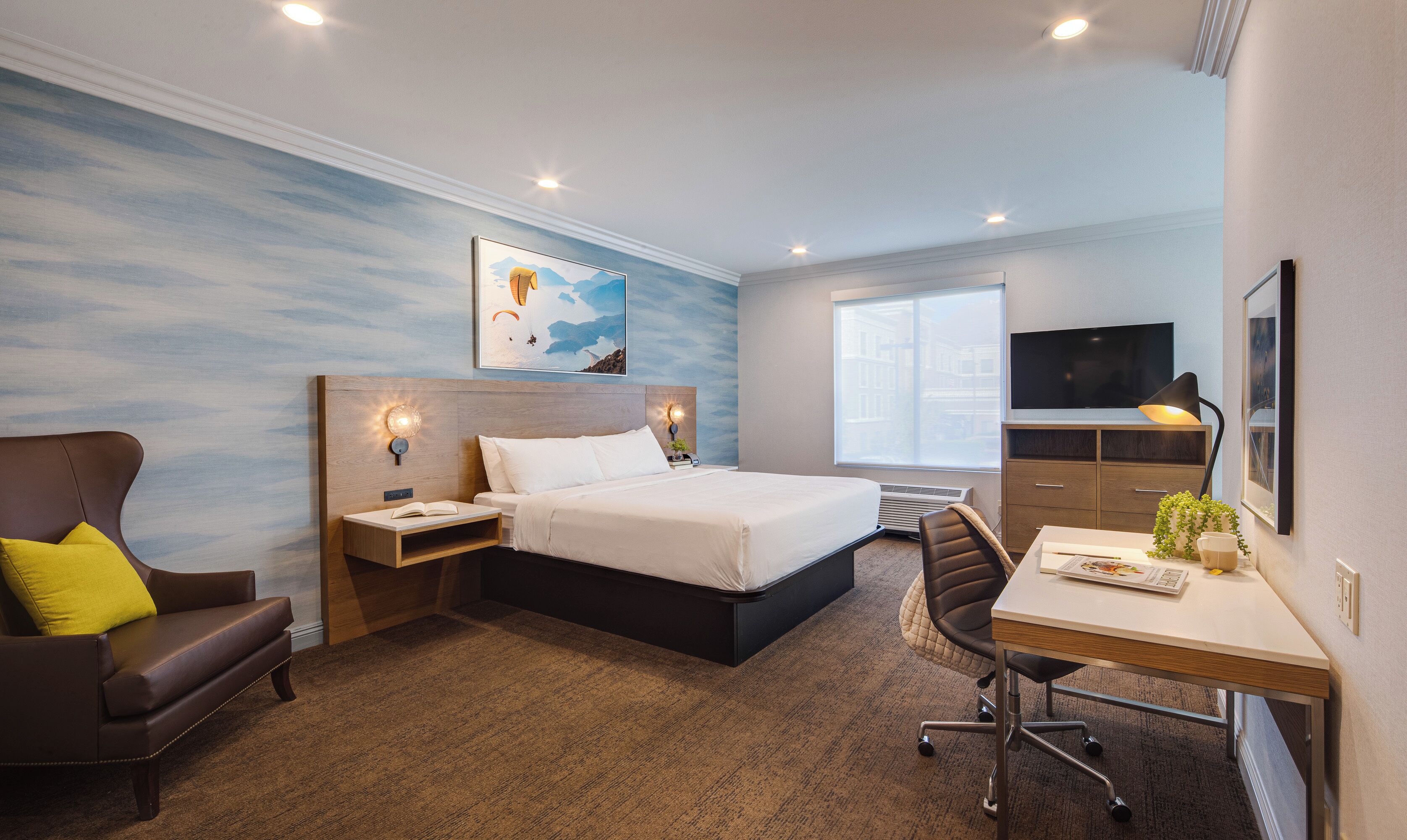 A hotel room with a bed, a desk, a chair, a TV, and a painting on the wall.
