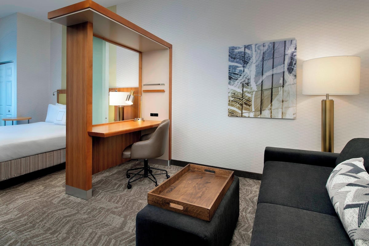 A hotel room with a bed, a desk with a chair, a sofa, a tray table, a lamp, and a painting on the wall.