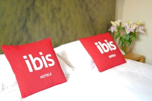 A hotel bed with two red pillows labeled "ibis HOTELS" and a vase of flowers on the nightstand.
