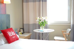 A hotel room with a bed, a red pillow, a small table with a vase of flowers, a chair, and a window with curtains.