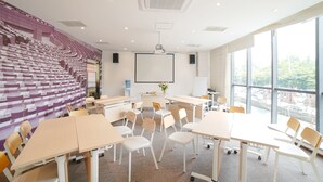 Un salón de conferencias con escenario, mesas y sillas blancas, ventanas amplias y una pantalla para proyector.