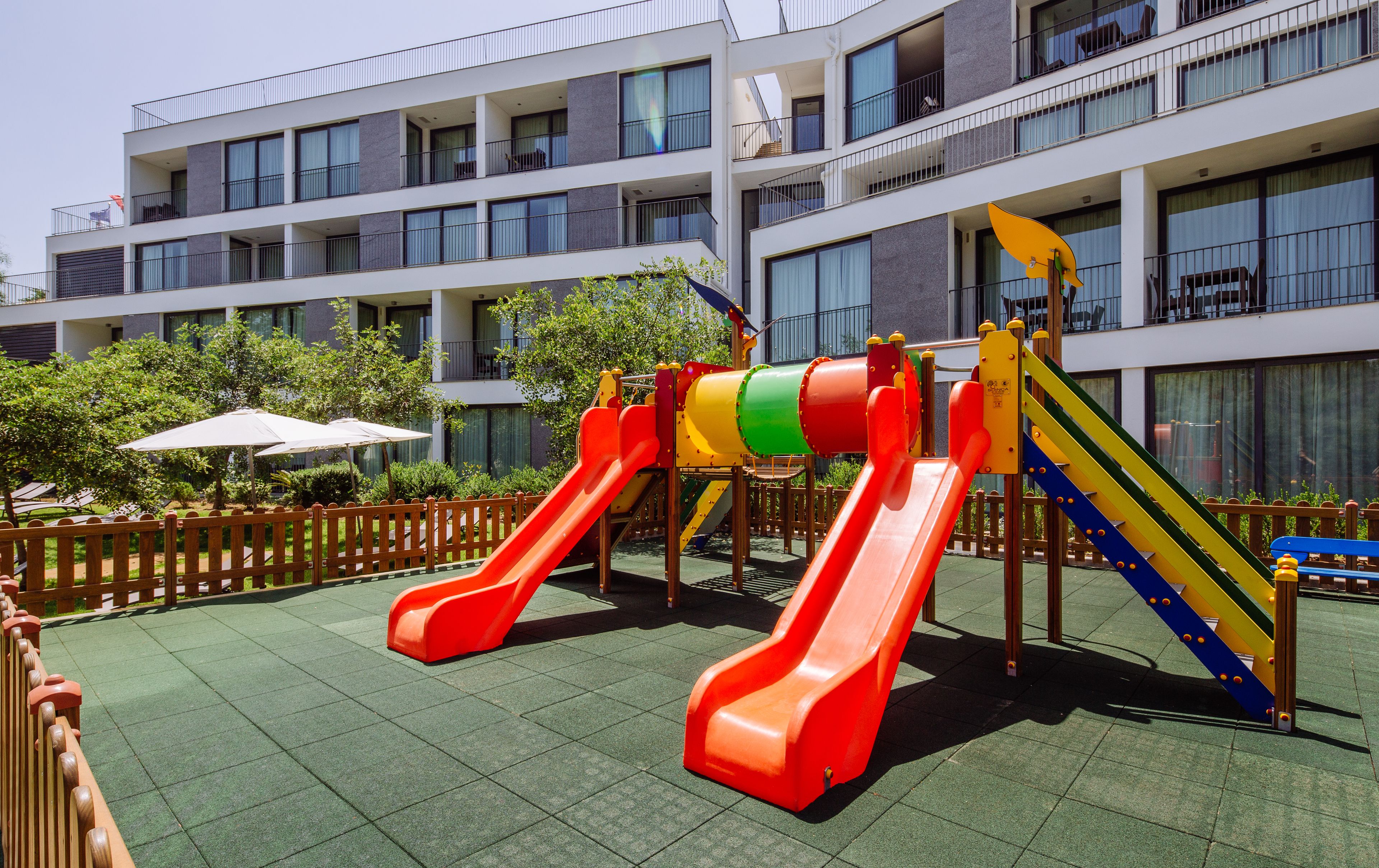 A playground with slides and climbing structures, surrounded by a wooden fence and greenery.