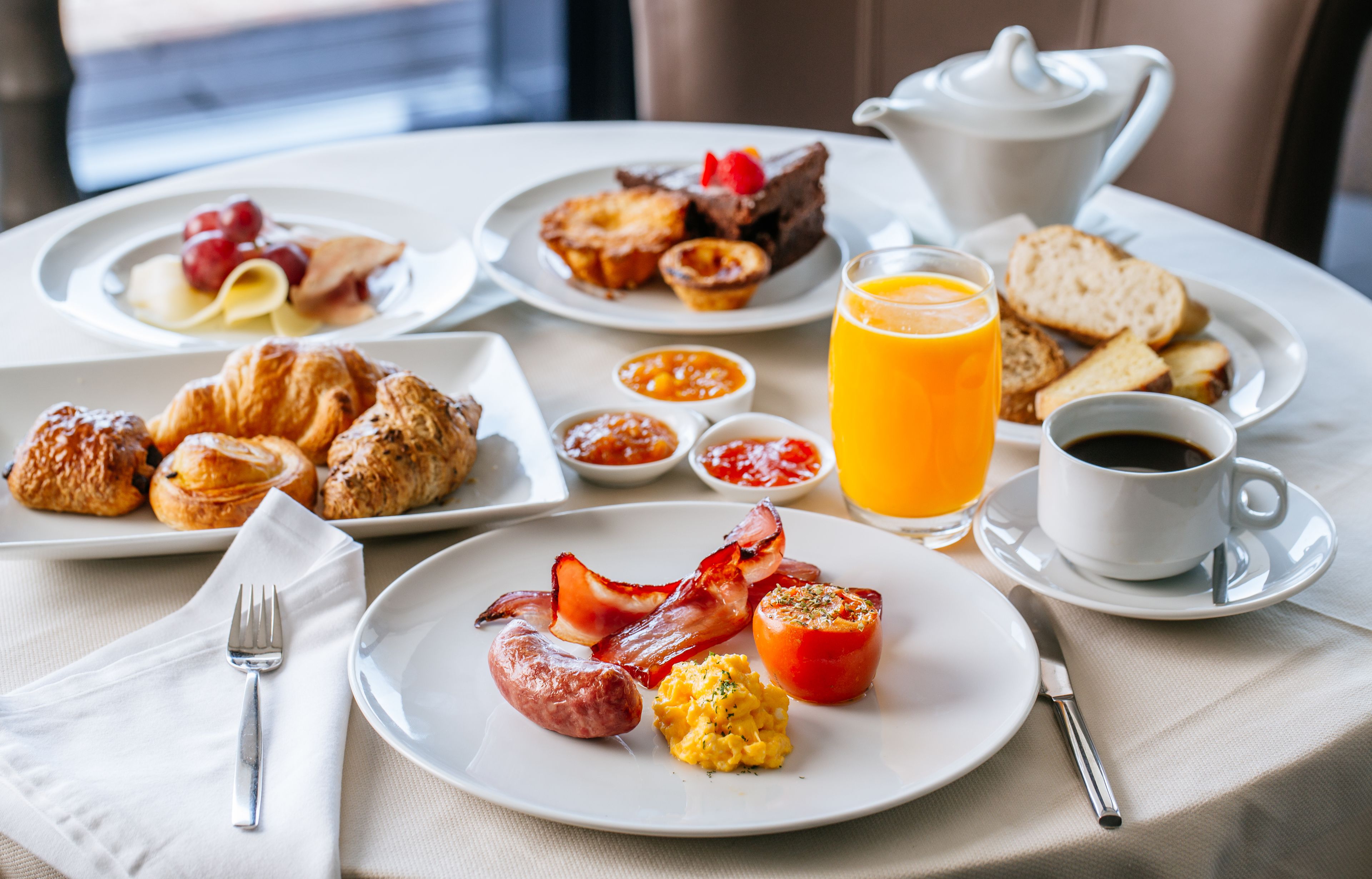A breakfast table with plates of eggs, sausages, and pastries, a glass of orange juice, and a cup of coffee.