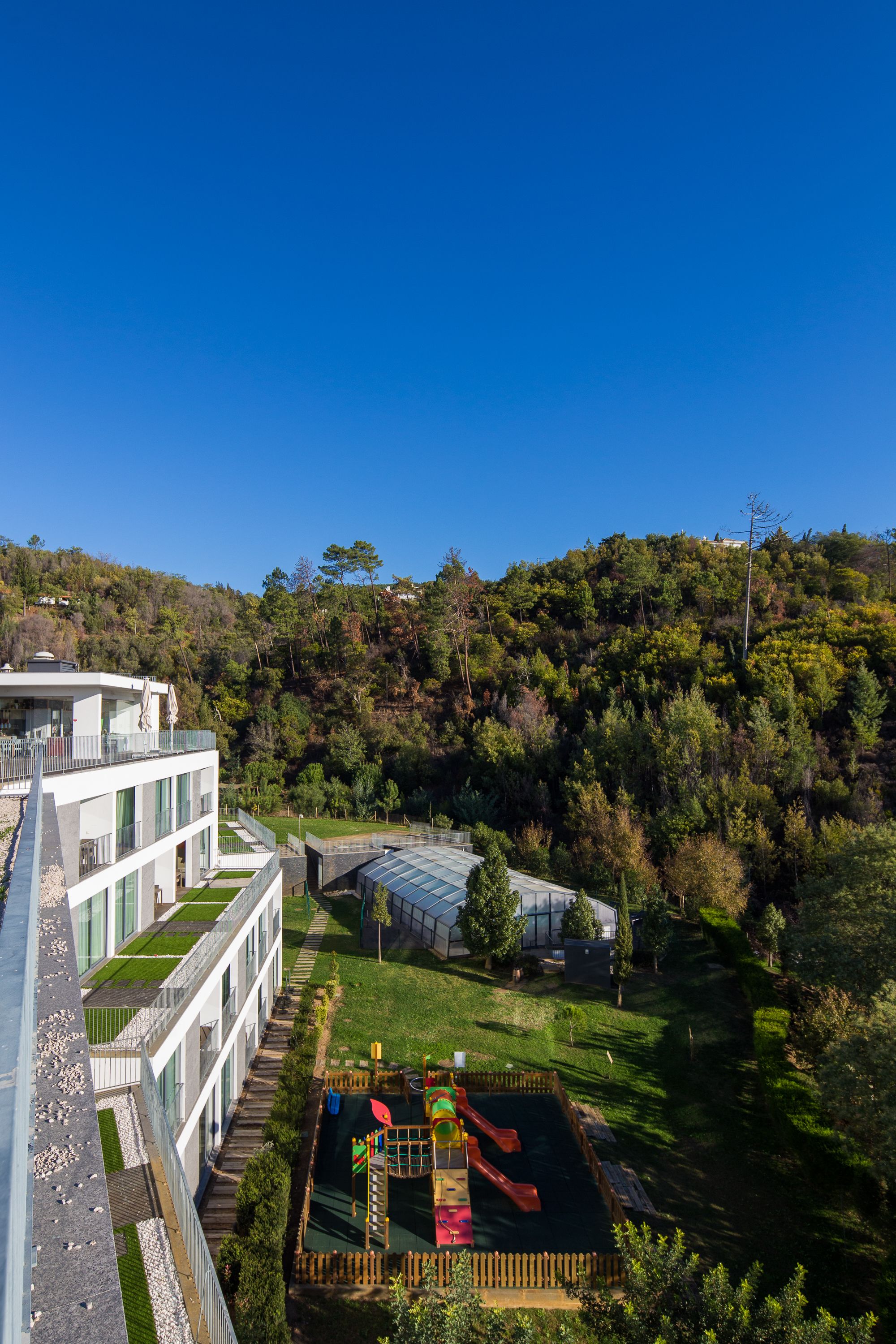 A modern building with a flat roof and large windows, surrounded by a lush green landscape and a playground.