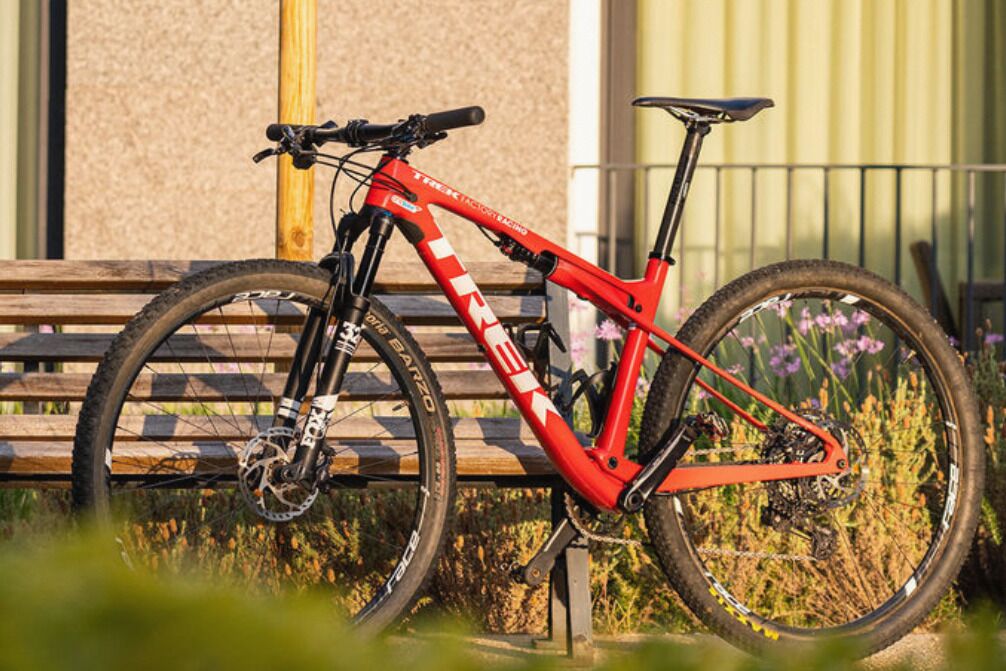 Una bicicleta de montaña roja estacionada en un banco de madera con una barandilla de metal al fondo.