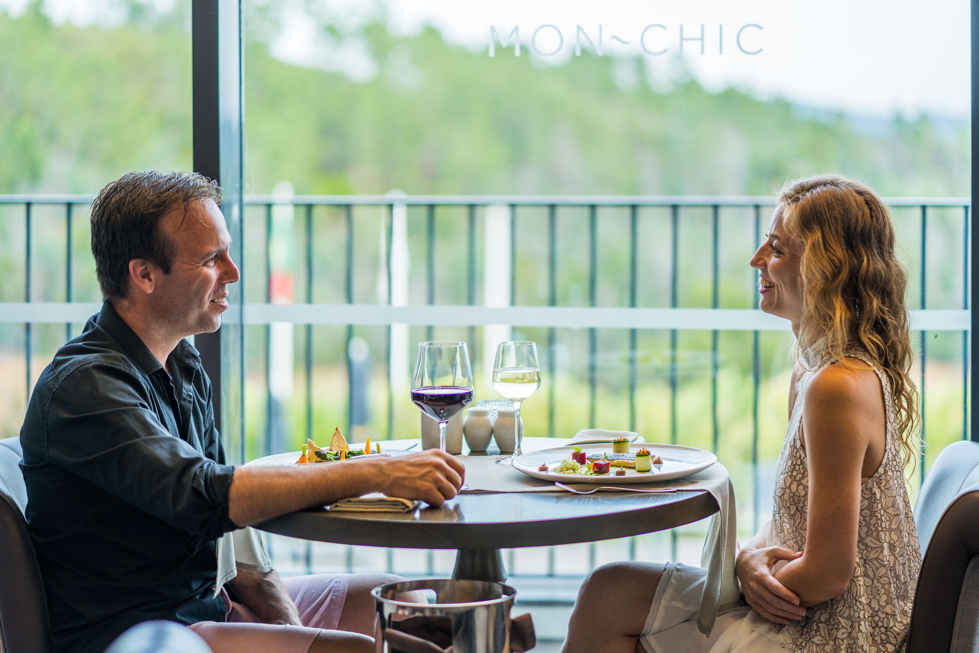A dining table with a view of the outdoors, two people seated, and a bottle of wine.