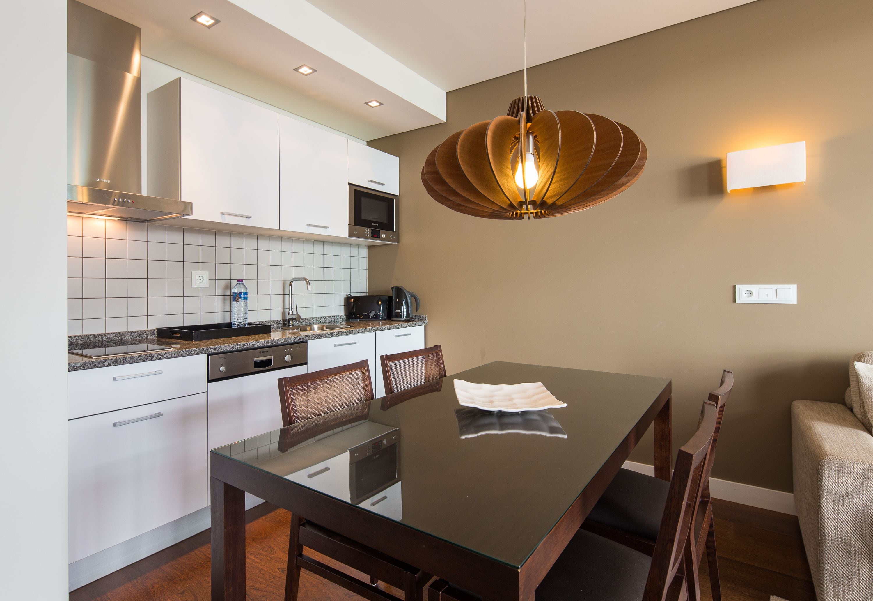 A modern kitchen with white cabinets, a microwave, and a coffee maker. There is a dining table with chairs and a pendant light above it.