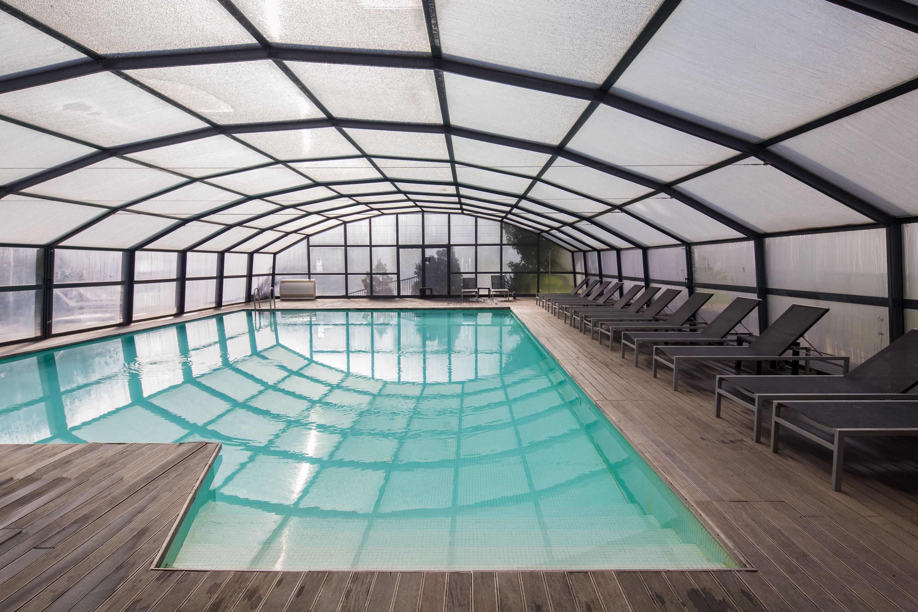 An indoor swimming pool with a glass ceiling, wooden deck, and lounge chairs along the sides.