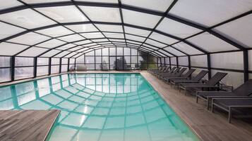 An indoor swimming pool with a glass ceiling, wooden deck, and lounge chairs along the sides.