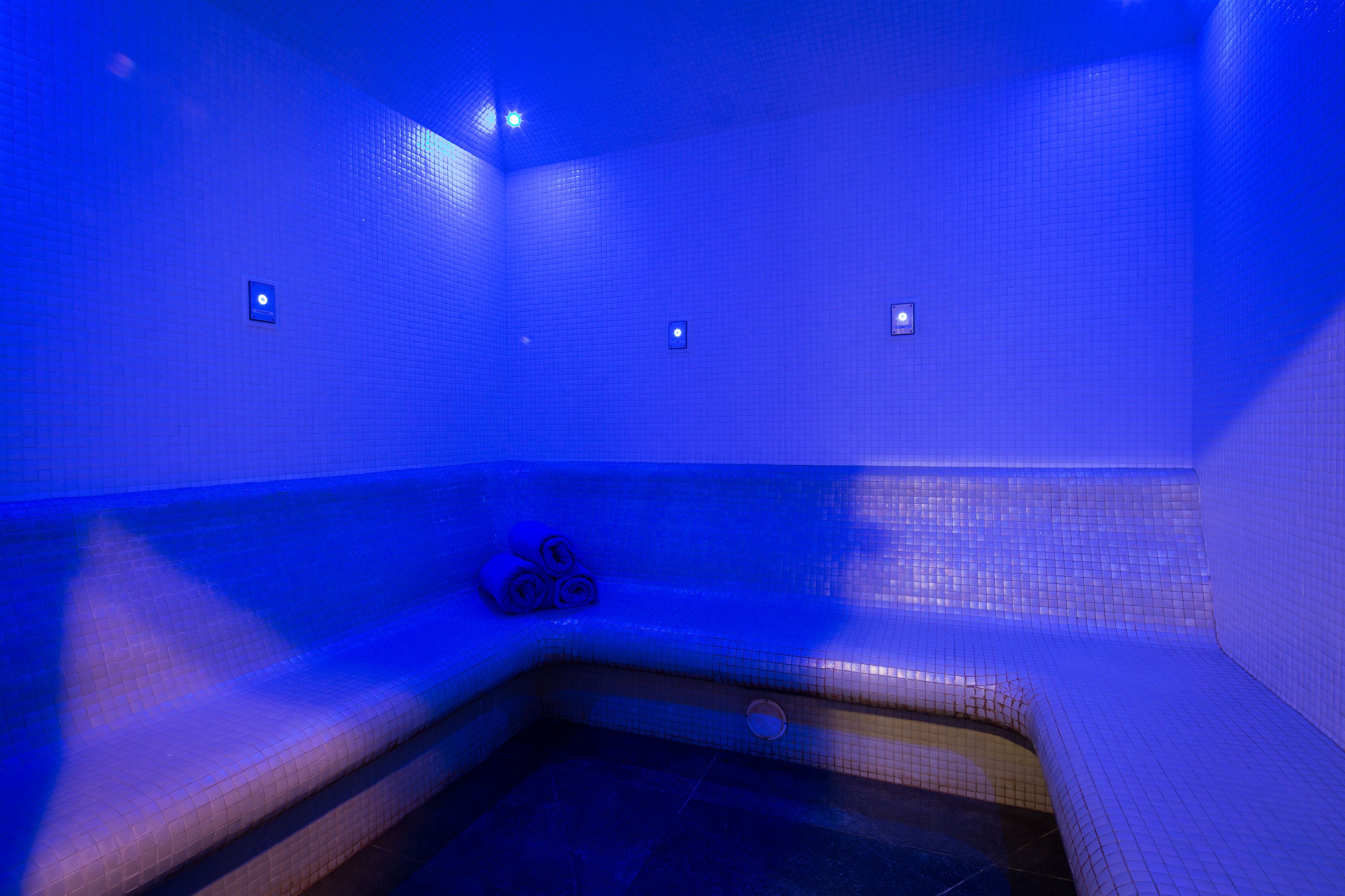Una habitación iluminada en azul con una bañera en esquina, una ventana pequeña y un perchero para toallas.