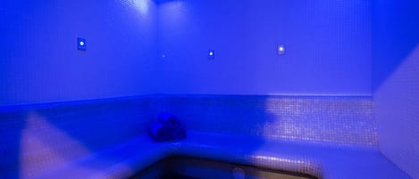 Una habitación iluminada en azul con una bañera en esquina, una ventana pequeña y un perchero para toallas.