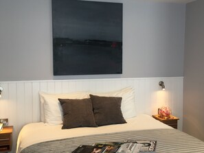Una cama bien hecha con almohadas blancas y marrones, una revista sobre la cama y un cuadro abstracto grande en la pared.