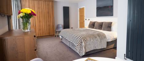 A modern bedroom with a bed, a wooden wardrobe, a desk with a chair, and a vase of flowers.