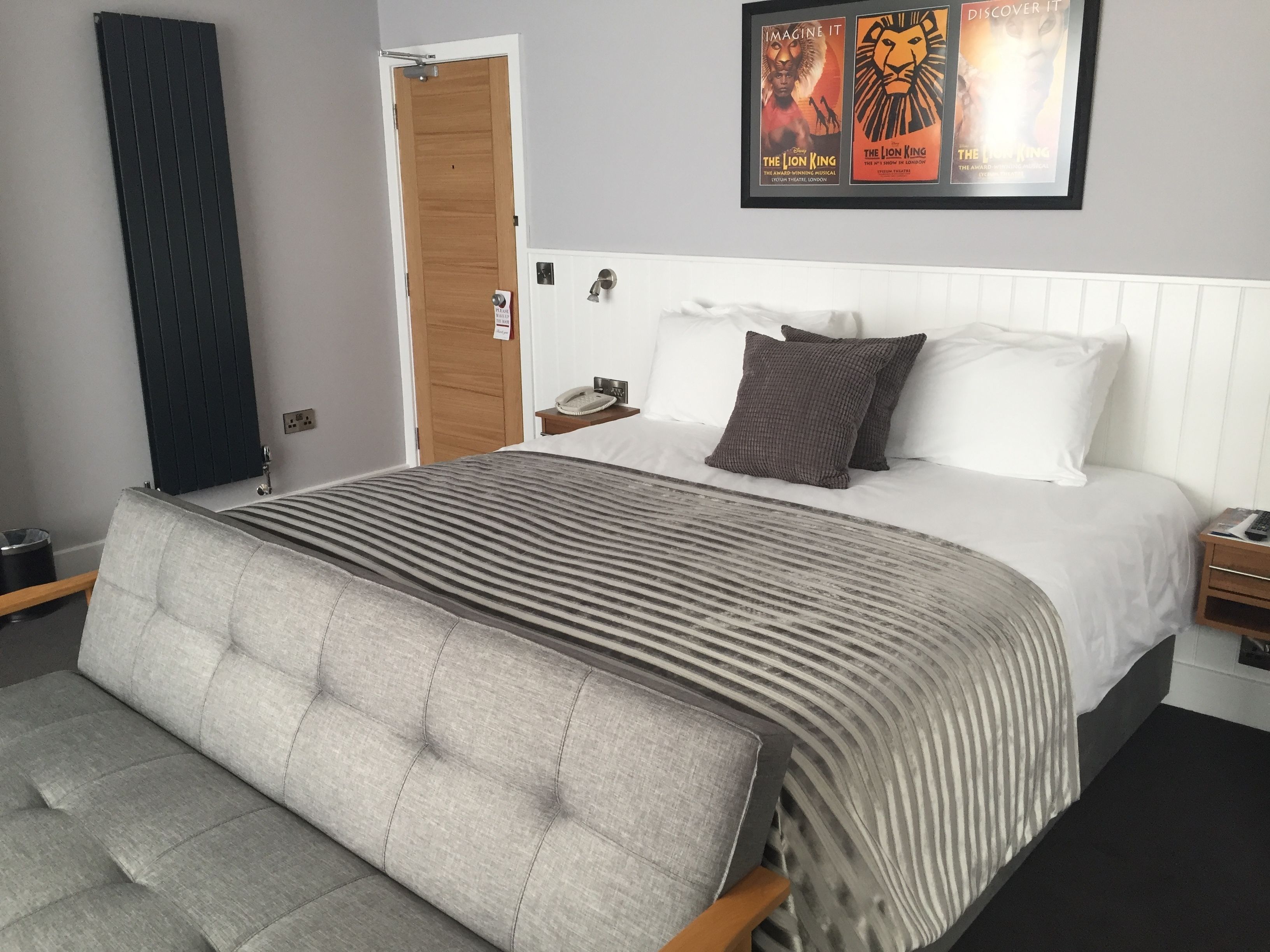 A bedroom with a bed, a bedside table, a nightstand, a framed poster on the wall, and a door.