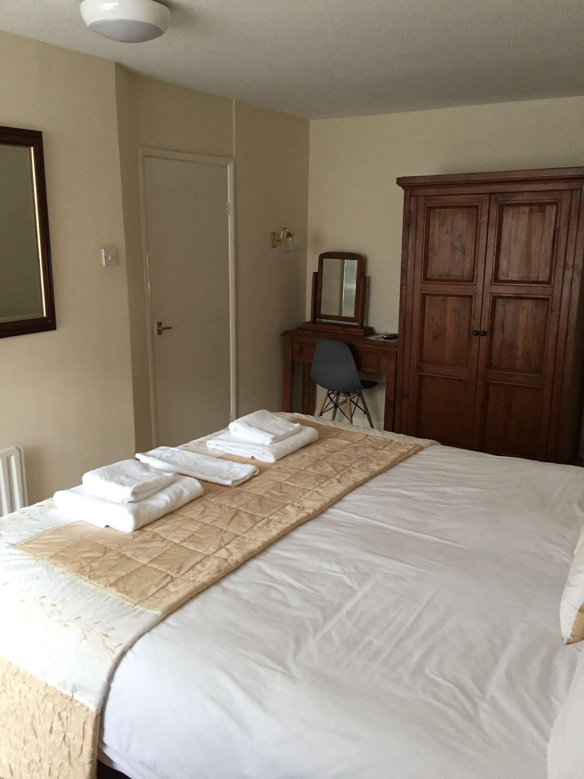 A neatly made bed with a quilt and folded towels, a wooden wardrobe, a desk with a chair, and a mirror on the wall.