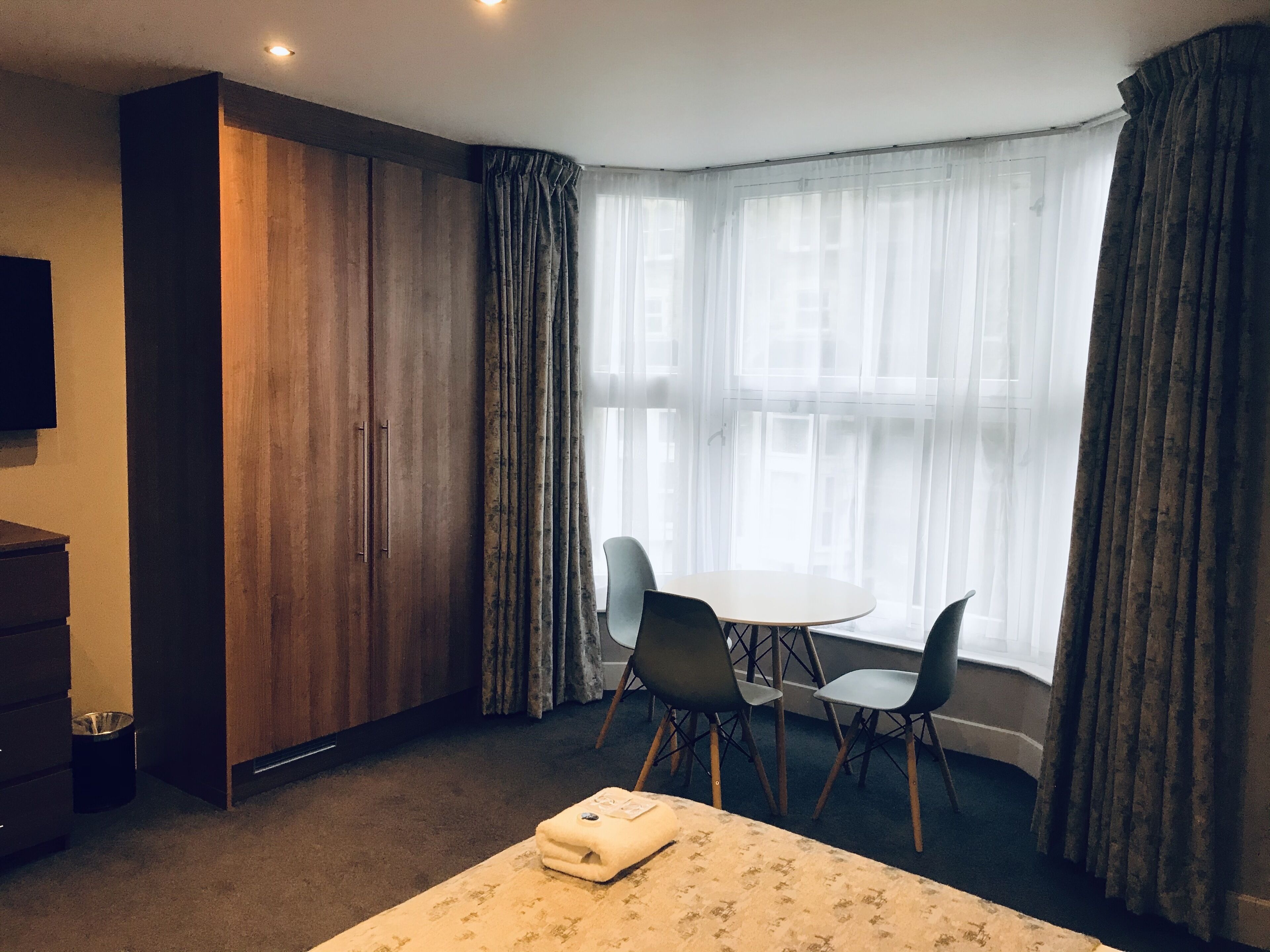 Ett hotellrum med en säng, ett skrivbord, två stolar, en garderob och ett stort fönster med gardiner.