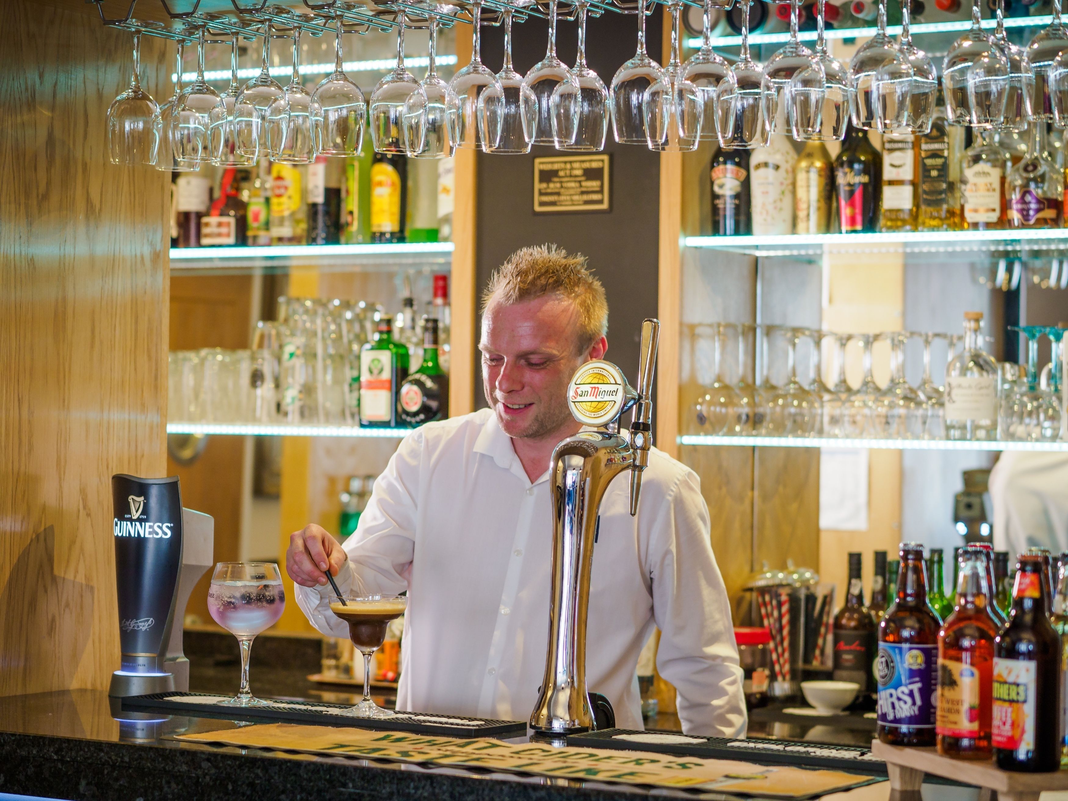 Ein Barkeeper zapft ein Getränk aus einem Zapfhahn mit der Aufschrift 'San Miguel' in einer Bar mit Regalen voller Spirituosenflaschen und Gläsern.