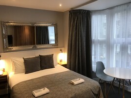 Una habitación de hotel con una cama grande, una mesita de noche con lámpara, una silla, una mesa redonda y un espejo grande sobre la cama.