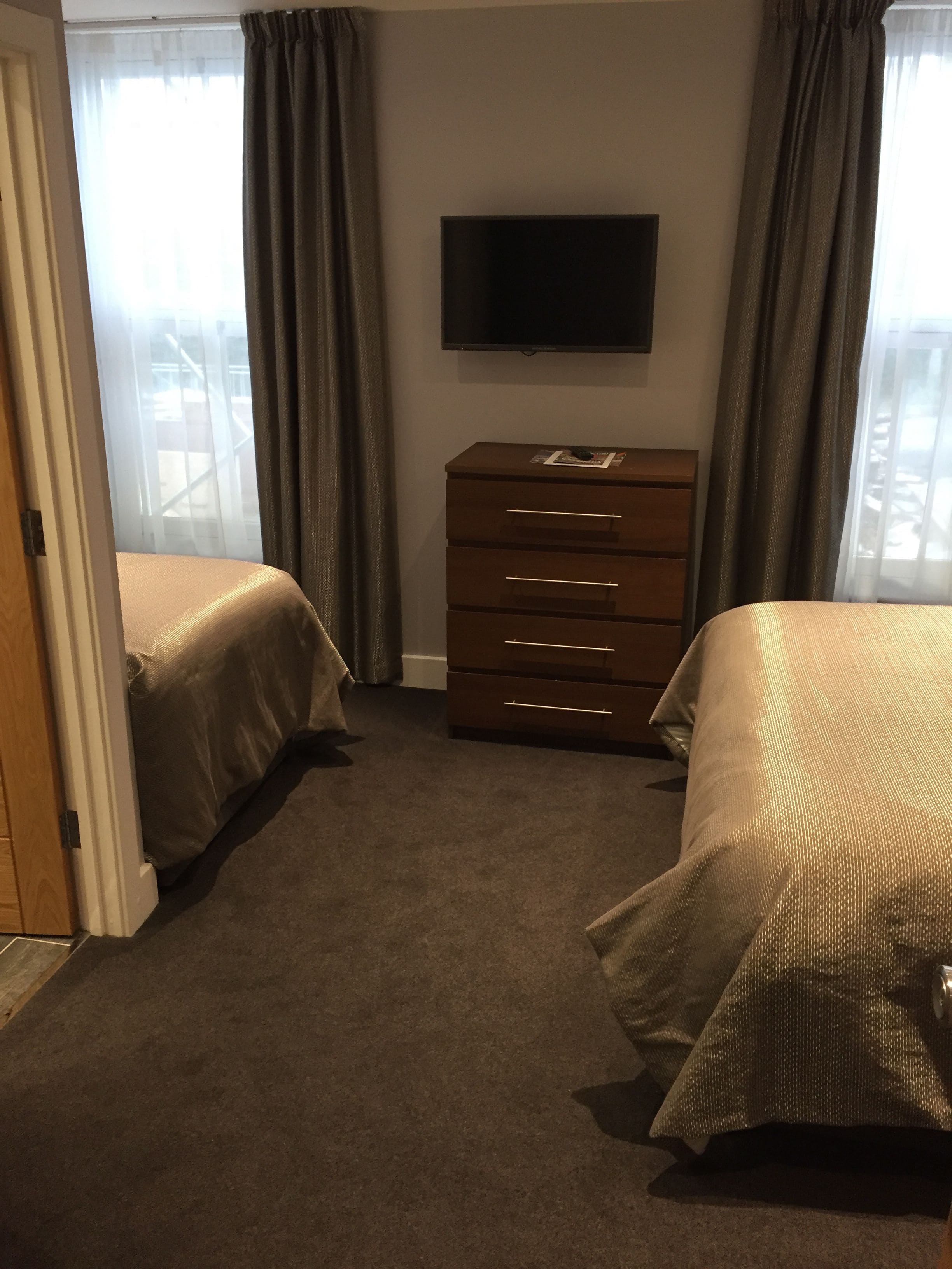 A hotel room with two beds, a dresser, a television on the wall, and a view of the outside through sheer curtains.