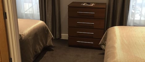 A hotel room with two beds, a dresser, a television on the wall, and a view of the outside through sheer curtains.