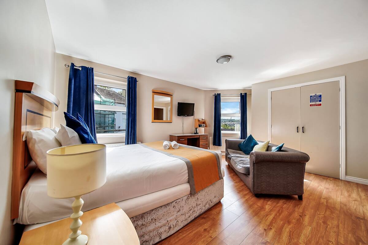A hotel room with a bed, a sofa, a desk, a TV, and a window with curtains.