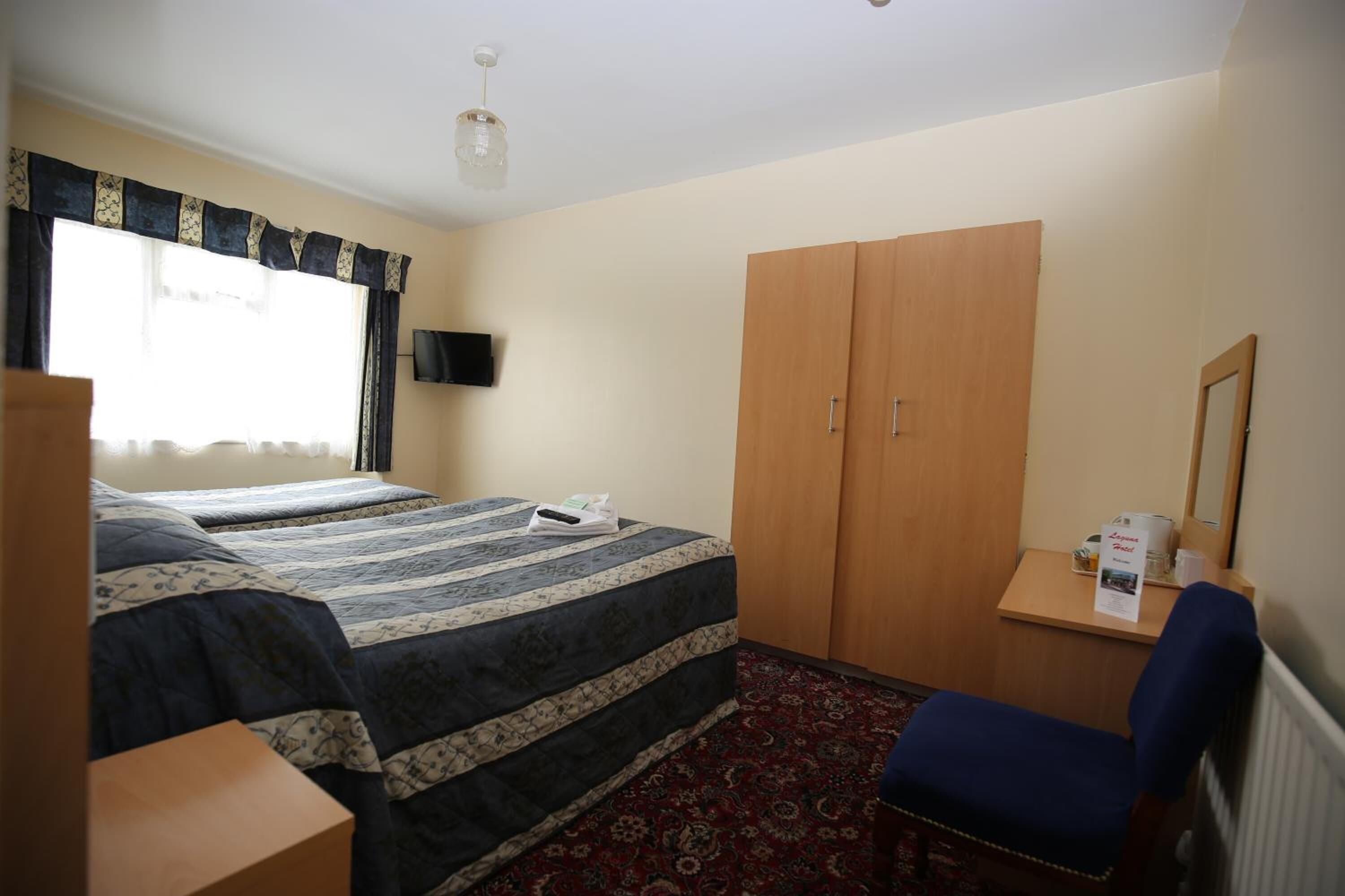 Une chambre d’hôtel avec deux lits, une armoire en bois, un bureau avec une chaise, une télévision et une fenêtre avec des rideaux.