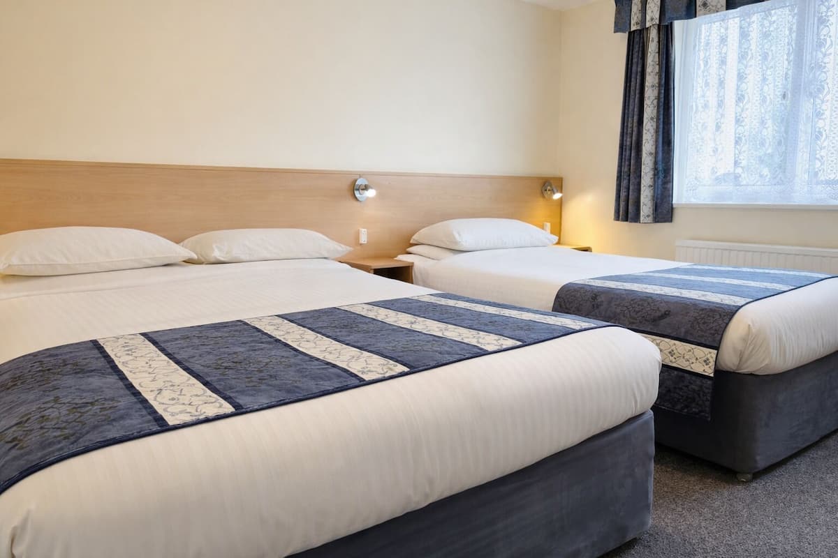 A hotel room with two beds, each with a blue and white quilt, white pillows, and a wooden headboard. There is a nightstand with a lamp between the beds, and a window with curtains.