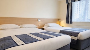 Quarto de hotel com duas camas, cada uma com colcha azul e branca, travesseiros brancos e cabeceira de madeira. Há uma mesinha de cabeceira com abajur entre as camas, e uma janela com cortinas.