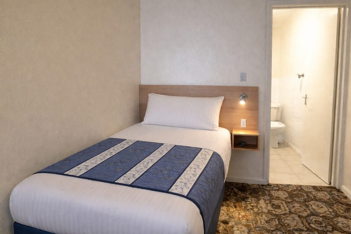 A single bed with a patterned bedspread, a bedside table with a lamp, and a bathroom visible through an open door.