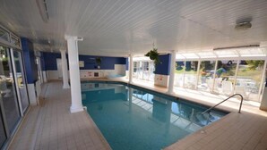 Piscina interior com paredes de vidro transparente, chão revestido e área ao lado da piscina com espreguiçadeiras e bar.