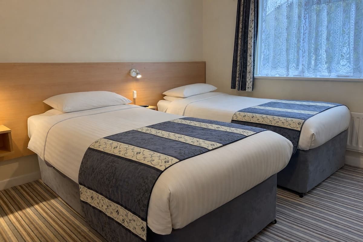 A hotel room with two beds, each with a patterned bedspread, a bedside lamp, and a window with curtains.